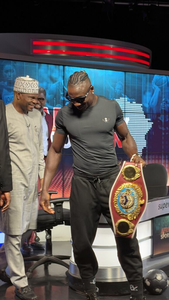 Zodsolo, in an AfroAlien t-shirt, looks down at his WBO Africa title belt in a television studio.
