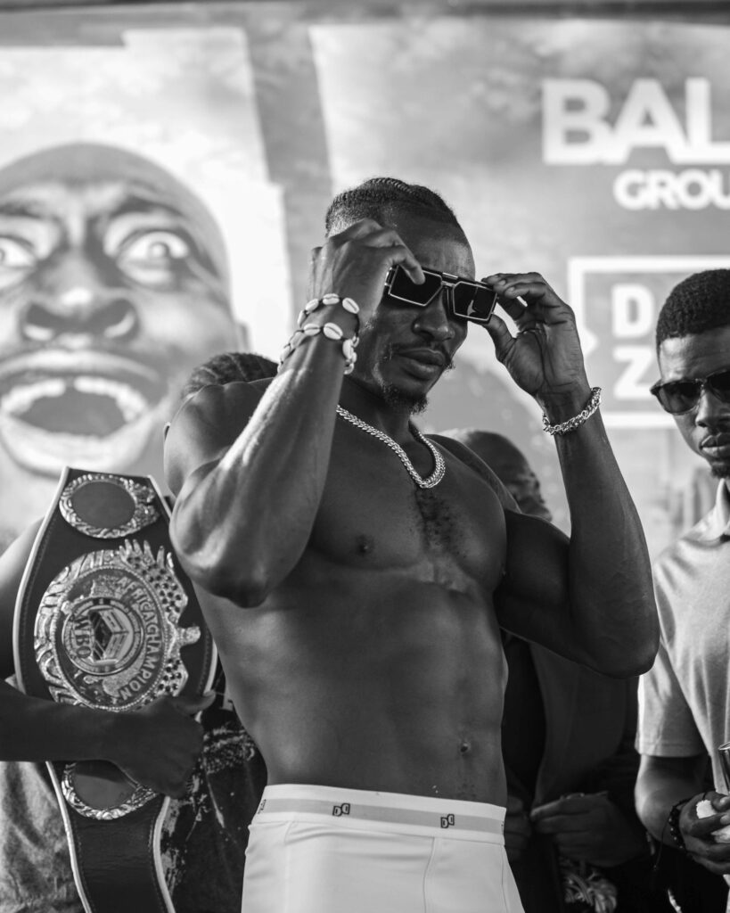 Black-and-white photo of Zodsolo adjusting his sunglasses beside the WBO Africa champion belt at a pre-fight event.